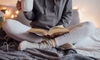 A woman sitting on a bed in sweat pants and a hoodie, holding a coffee mug and a book. 