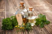 Two bottles of olive oil sit on a wooden surface with herbs surrounding them.