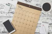 A January calendar on a wood table surrounded by a notepad, calculator, phone, and cup of coffee. 