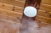 A smoke detector on a wood ceiling with smoke around it. 