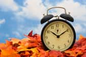 A clock sitting in a pile of fall leaves against a blue cloudy sky. 