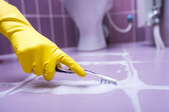 Gloved hand scrubbing grout between purple floor tiles in a bathroom