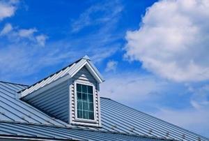 An attic window jutting out from a tin roof with clouds beyond.