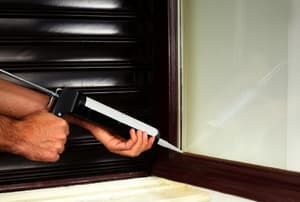 Hands holding a caulking gun up to a window from inside.