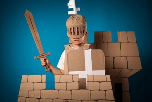 A boy playing in a cardboard castle with a cardboard sword and helmet.