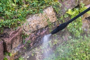 hand with pressure washing wand spraying stone wall with greenery on it