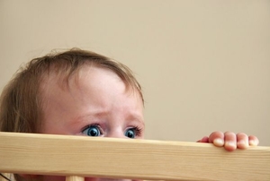 A baby looking over the baby gate with a scared look in her eyes.