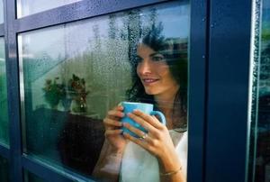 A woman holding a mug with two hands and looking out a rain-covered window.