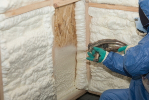 person in blue protective gear spraying white foam insulation on wooden walls