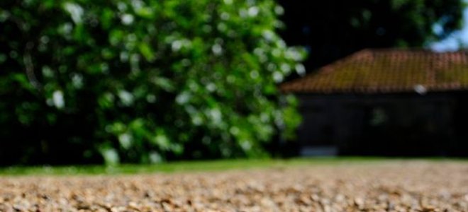 A close-up image of a gravel driveway with a house and a tree in the background.