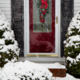 Christmas wreath on the front door of a house, surrounded by snow-covered bushes.