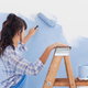 A woman stands on a ladder to roll light blue paint onto a white wall.