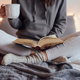 A woman sitting on a bed in sweat pants and a hoodie, holding a coffee mug and a book. 
