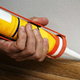 Using a caulking gun, sealant is applied along the edge of a piece of baseboard trim.