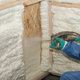 person in blue protective gear spraying white foam insulation on wooden walls