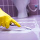 Gloved hand scrubbing grout between purple floor tiles in a bathroom
