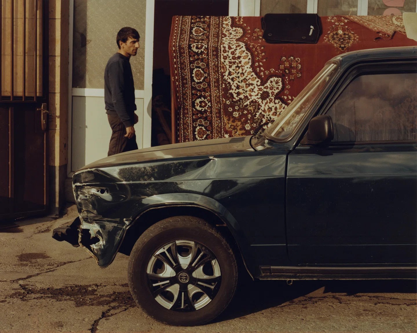 Front end of a bullet-riddled Lada Car parked in front of a striking Oriental rug.