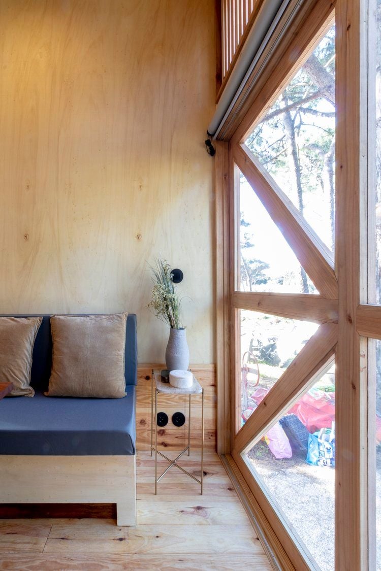 Small wooden couch and end table tucked into a corner of the Adraga House tiny home near a large glass pane.
