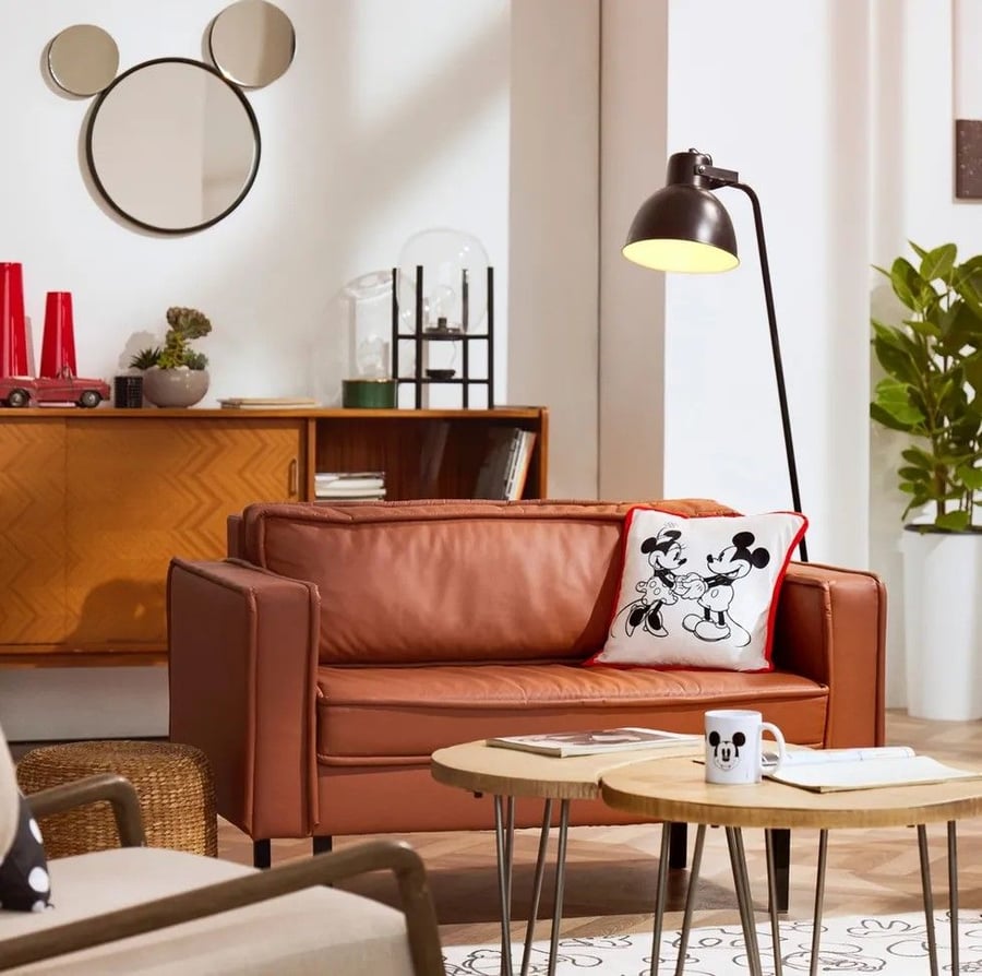 Living space filled with splashes of fun in the form of Disney Home decor pieces, from a mug and throw pillows to a mouse-shaped mirror on the wall.