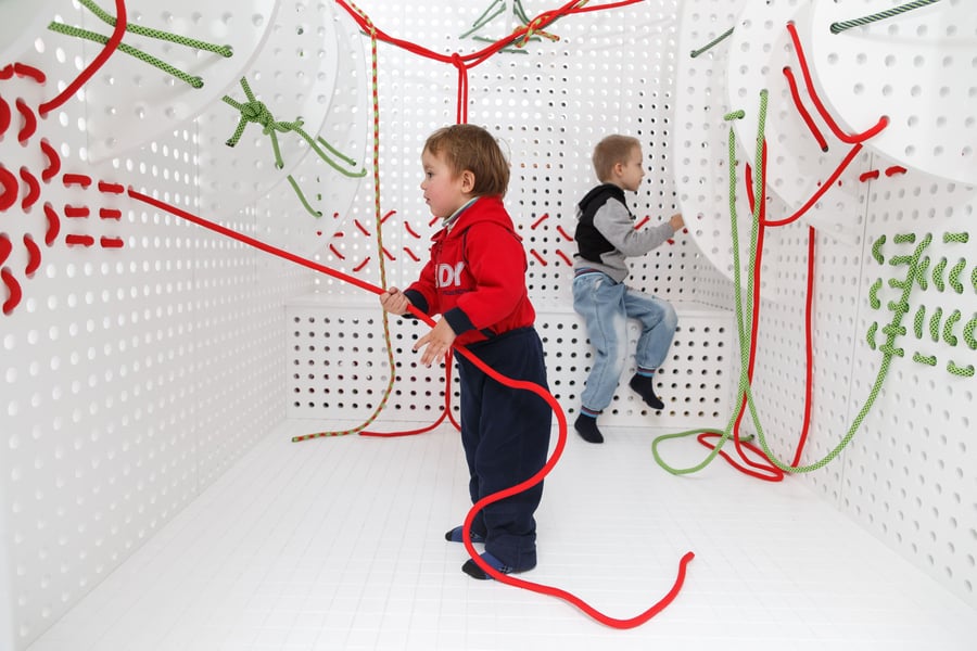 Two kids use colorful cords to stitch patterns in the walls and seats of the all-white CUBA BUBA Matrix module. 