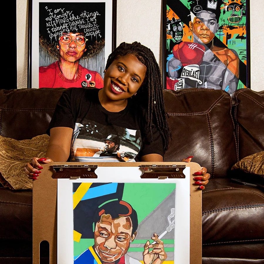 Artist Domonique Brown holds up a portrait of the writer James Baldwin, beaming with pride in front of two other pieces from her DomoINK Art brands.