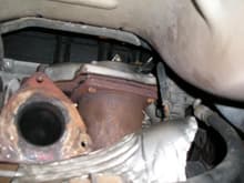 Despite what the service manual says, you cannot get the Y-pipe out of the car at this point.  The large three-bolt flange will not pass by the rear catalytic converter and the rear engine mount.  In this picture you can see the flange at the left and the rear cat to its right.  Notice the bolts holding the cat.  They do not turn.  There are nuts at the top of the bolts.  You must remove these nuts to remove the cat.  There are four holding the cat to the exhaust manifold.  You will be able to see the rear two nuts to remove them, but you must do the front two nuts entirely by feel.  Use a high quality 13 mm socket.  Use a short extension for the right front nut to lift the wrench above the rear O2 Sensor, the top of which is visible in the picture.  Theres just enough room to do this, working from the rear over the crossmember.