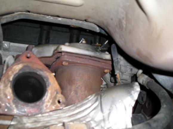 Despite what the service manual says, you cannot get the Y-pipe out of the car at this point.  The large three-bolt flange will not pass by the rear catalytic converter and the rear engine mount.  In this picture you can see the flange at the left and the rear cat to its right.  Notice the bolts holding the cat.  They do not turn.  There are nuts at the top of the bolts.  You must remove these nuts to remove the cat.  There are four holding the cat to the exhaust manifold.  You will be able to see the rear two nuts to remove them, but you must do the front two nuts entirely by feel.  Use a high quality 13 mm socket.  Use a short extension for the right front nut to lift the wrench above the rear O2 Sensor, the top of which is visible in the picture.  Theres just enough room to do this, working from the rear over the crossmember.