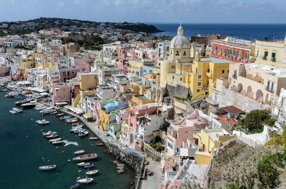 The iconic belvedere vista of Corricella. Amid the pastel buildings, yellow chiesa S. Rocco looms large. Nearby, S.M. delle Grazie, Casale Principe Umberto and Palazzo Mignano de Iorio. 